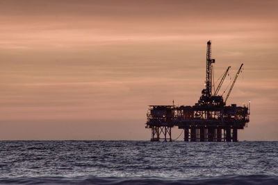 Silhouette cranes oilplatform  by sea against sky during sunset