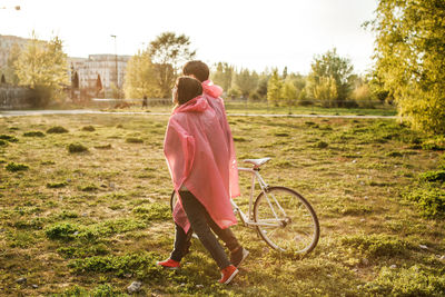 Young couple in pink raincoat walking with bicycle on field against sky
