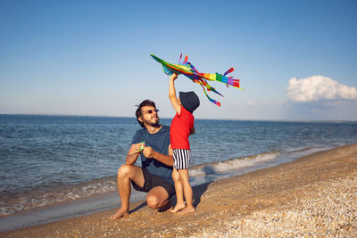 Father and son are standing on a sandy beach by the sea and launch a toy striped kite in the summer