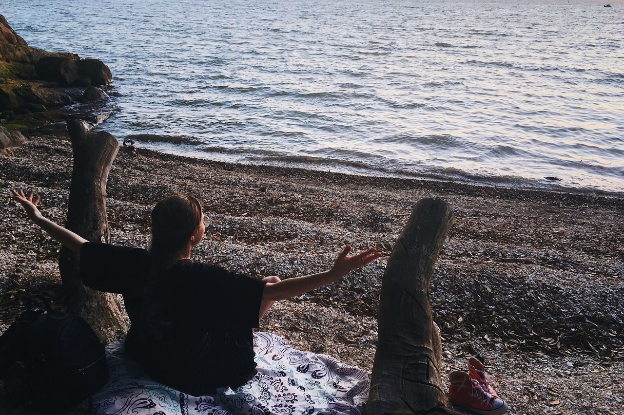 REAR VIEW OF WOMAN SITTING ON ROCK AT SHORE