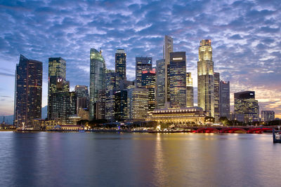 Illuminated modern buildings by river against sky