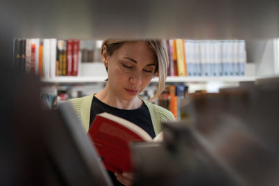 Female mature student looking for book at library bookshelf studying at university campus or college