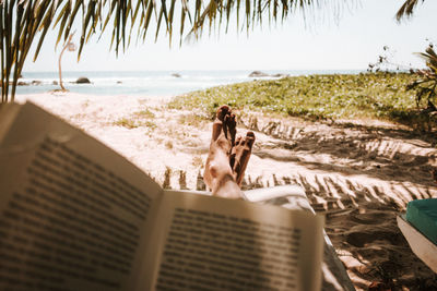 Low section of man reading book on beach during sunny day