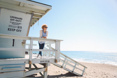 Woman leaning on railing of lifeguard hut at beach against clear sky