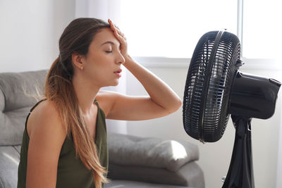 Overheated young woman in front of the working fan suffering from summer heat at home.