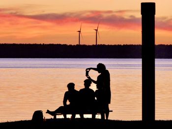 Silhouette men sitting on shore against orange sunset sky