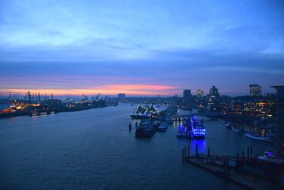 Illuminated city by river against sky at sunset