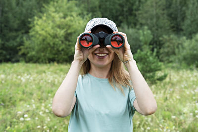 Happy young blonde woman bird watcher in cap looking through binoculars at cloudy sky in forest 