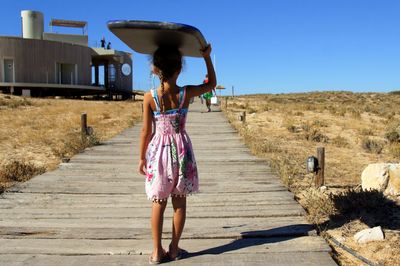 Rear view of girl standing with surfboard on head over boardwalk at beach