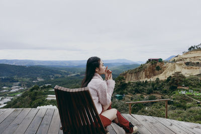 Side view of woman sitting on retaining wall against sky