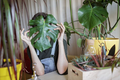 A young girl covers her face with green leaf for fun and relax, home plants on the balcony, gen z