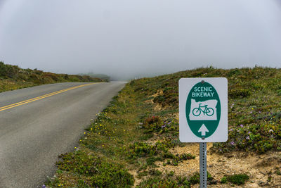 Road sign on field by empty road against cloudy sky