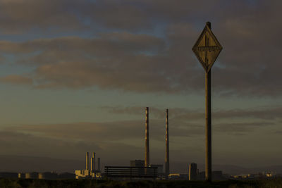 Pole and smoke stacks against cloudy sky at sunset