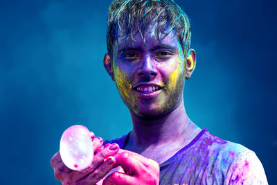 Portrait of young boy holding water balloon with face covered in colors against blue background
