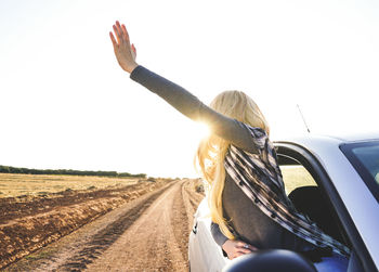 Young woman with arms outstretched leaning out from car window against clear sky