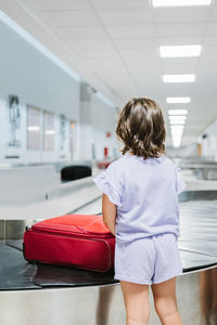 Back view of unrecognizable girl in lilac clothes waiting for luggage to arrive on carousel in airport terminal during trip