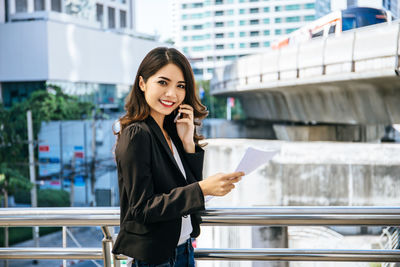 Portrait of businesswoman talking on phone while holding document by railing on bridge