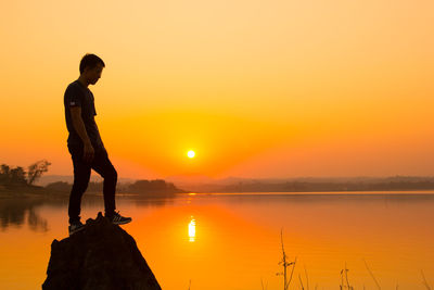 Full length of young man on rock by lake against sky during sunset