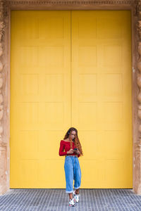 African american female standing near huge wooden doors while on entrance of building with mobile phone listening to music on headphones