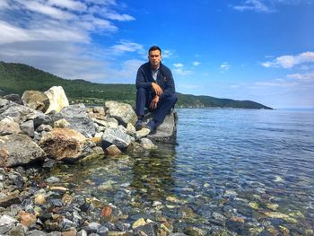 Portrait of young man sitting on rock at lake against sky