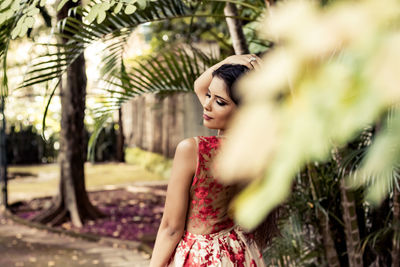 Woman looking away while standing with hand in hair by trees