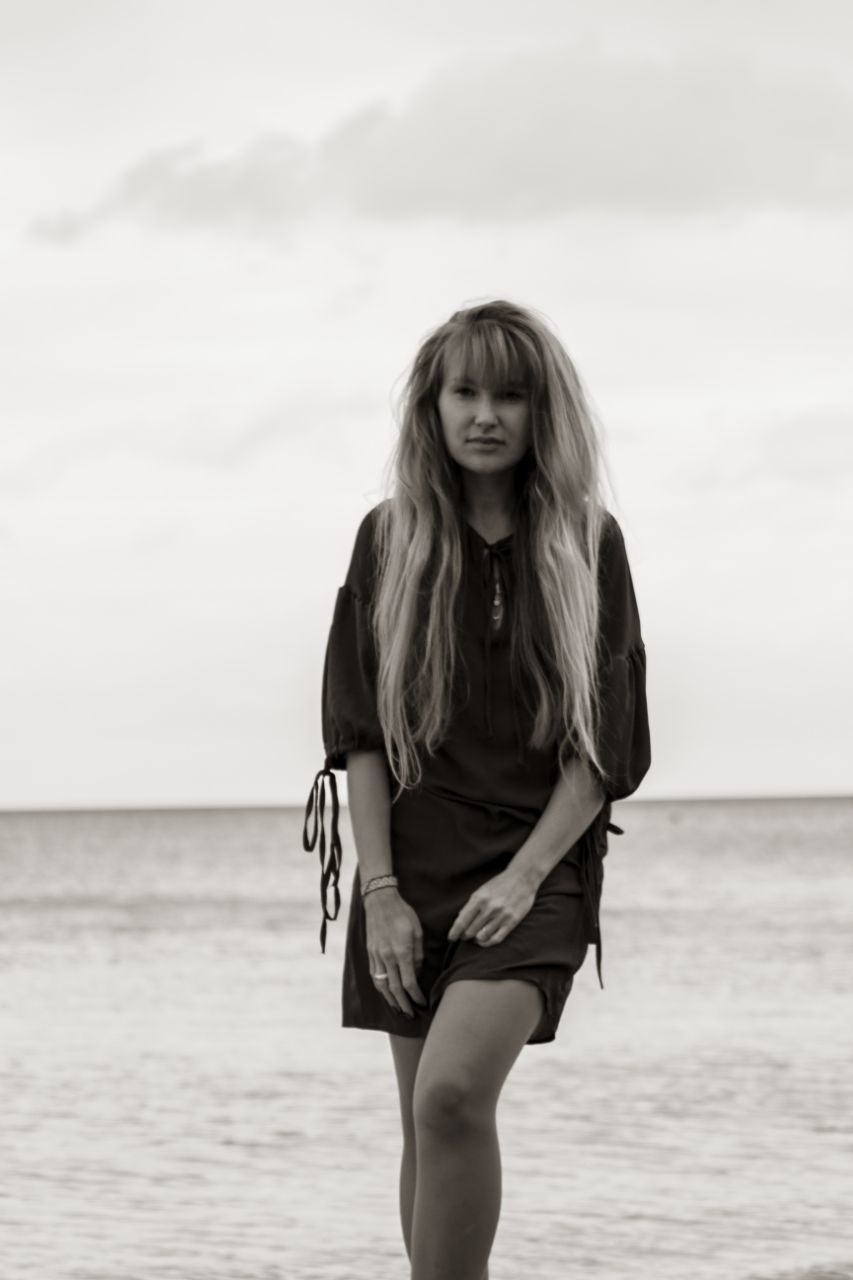 one person, long hair, sea, women, hairstyle, beach, water, female, land, sky, white, young adult, adult, black, dress, clothing, photo shoot, fashion, nature, portrait, black and white, horizon over water, leisure activity, front view, horizon, portrait photography, monochrome photography, standing, looking at camera, lifestyles, day, person, emotion, monochrome, holiday, smiling, three quarter length, blond hair, beauty in nature, casual clothing, cloud, outdoors, trip, summer, happiness, vacation, sand, full length, human hair, sitting, human leg, spring