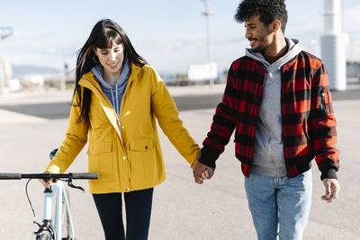 Smiling woman with bicycle holding hand of male friend while walking on footpath