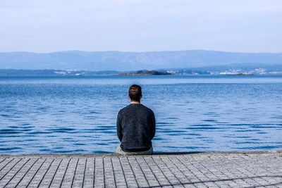 Rear view of man sitting on pier over sea against mountain