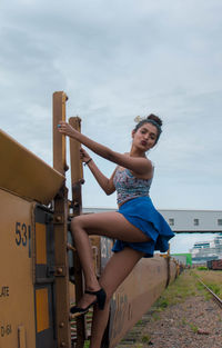 Portrait of young woman puckering lips while standing on ladder against cloudy sky