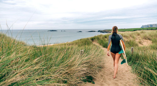 Surfer woman with wetsuit and surfboard walking along the path between the dunes towards the beach