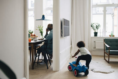 Girl riding toy car in living room while father having breakfast at dining table