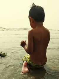 Rear view of shirtless boy at beach against sky