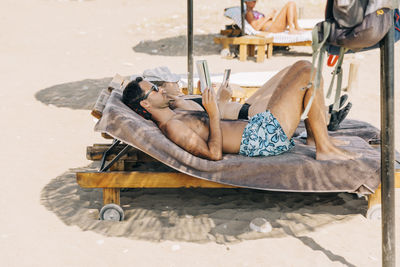 Shirtless man reading book while lying on lounge chair at beach