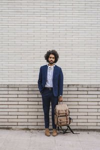 Male entrepreneur holding backpack while standing with hand in pocket on footpath