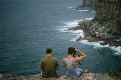 Rear view of male friends sitting on cliff against sea