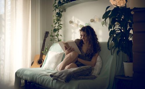 Young woman reading book while sitting on sofa at home