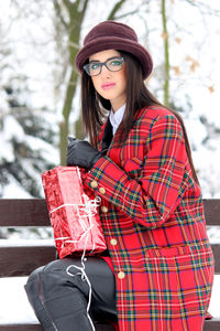 Portrait of young woman wearing hat sitting against trees during winter