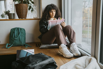 Full length of young woman writing in book while sitting by window at university