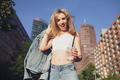 Low angle portrait of woman holding disposable cup against buildings