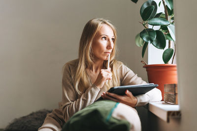 Beautiful blonde thinking young woman artist illustrator drawing on tablet near window at the home