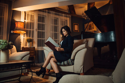 Side view of young woman reading book while sitting on armchair at home