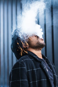 Side view of ethnic back guy with dreadlocks and hat in dense cloud of fume smoking on street in city