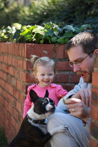 Smiling daughter looking at father playing with dog by brick wall in yard