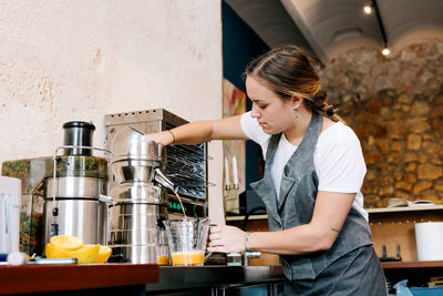 Side view of diligent barista preparing citrus fresh juice in shiny juicer at table in cafe