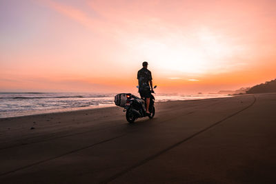 Man riding motorcycle on beach against sky during sunset