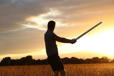 Man standing on field with sword against sky during sunset