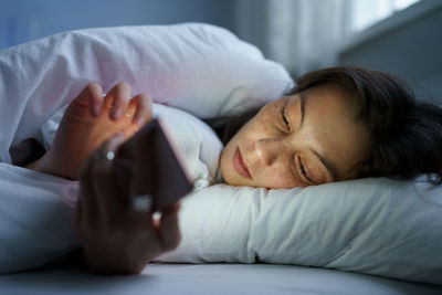 Young woman using mobile phone while lying in bed at night, suffering from post-breakup insomnia