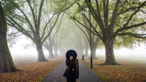 Woman covering face with hat while standing on road amidst trees during foggy weather