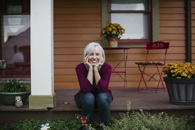 Portrait of cheerful senior woman with hands on chin sitting in porch