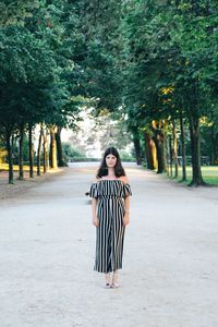 Full length portrait of young woman standing amidst trees on road